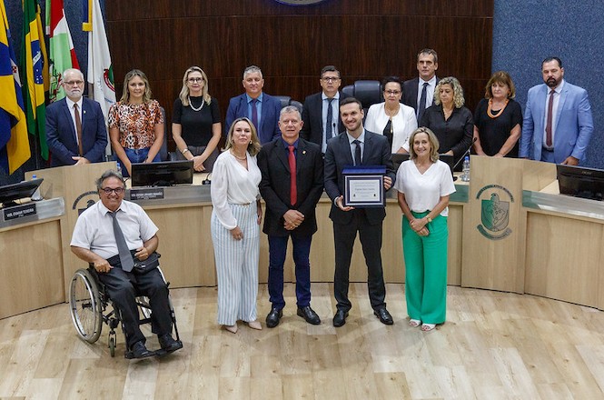 A imagem mostra um grupo de pessoas posando para uma foto em frente a uma bancada. No centro, um homem segura um certificado acompanhado de outras pessoas.