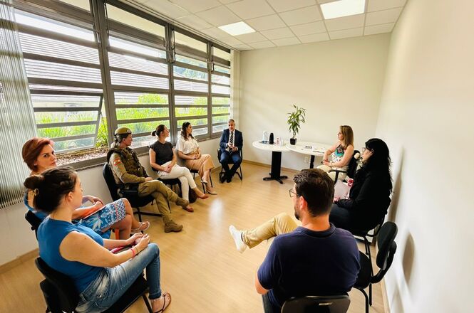 A imagem mostra um grupo de pessoas sentadas em cadeiras dispostas em círculo em um grupo reflexivo. A sala possui paredes brancas e piso de madeira clara.