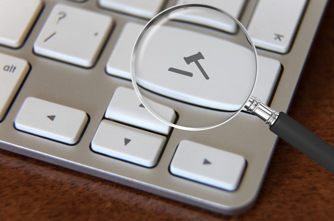 A imagem mostra o teclado de um computador com uma lupa aproximando uma das teclas. Na tecla está um malhete. O computador é branco e está em cima de uma mesa de madeira.