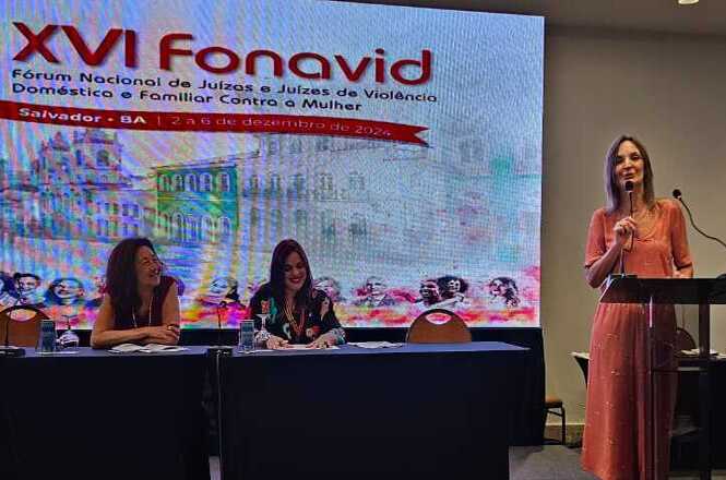 A imagem mostra um evento realizado no Fórum Nacional de Juízas e Juízes de Violência Doméstica e Familiar contra a Mulher (FONAVID). Em destaque, há três pessoas no palco: duas mulheres estão sentadas à mesa à esquerda, e outra está de pé, à direita, em um púlpito, discursando ao microfone.