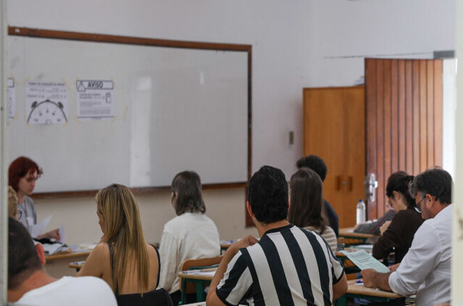 A imagem retrata uma sala de aula durante um concurso. Há várias pessoas sentadas em cadeiras com mesas e papeis à sua frente. No fundo, uma pessoa está sentada próxima a um quadro branco.