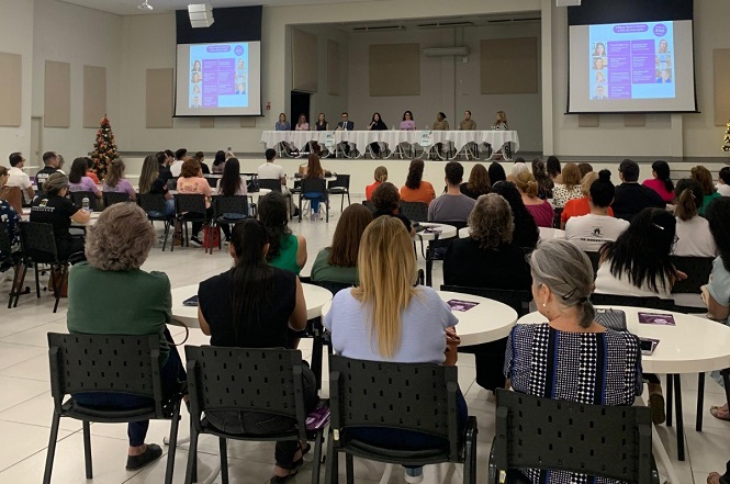 A imagem mostra um auditório onde ocorre o evento “Lei Maria da Penha em Debate”. No palco, há uma mesa composta por várias palestrantes, com microfones e telas grandes projetando informações. O público está sentado em mesas redondas.