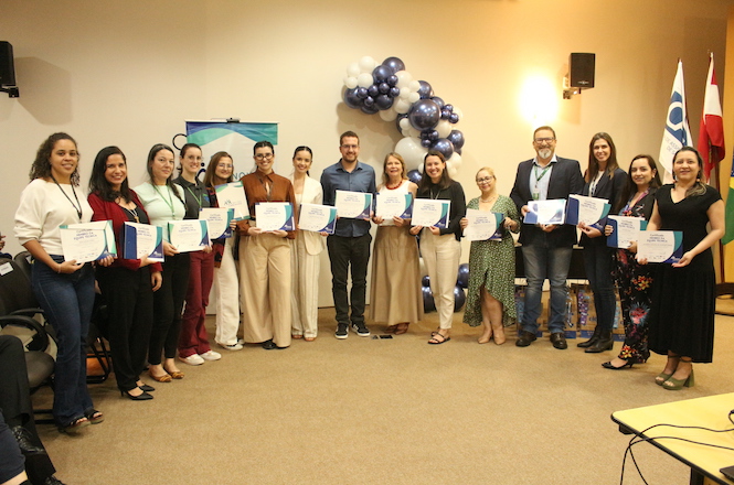 A imagem mostra um grupo de pessoas posando para uma foto, no evento do Programa Novos Caminhos. A maior parte das pessoas seguram certificados em mãos. Elas estão em um ambiente interno decorado com balões nas cores azul e branco.