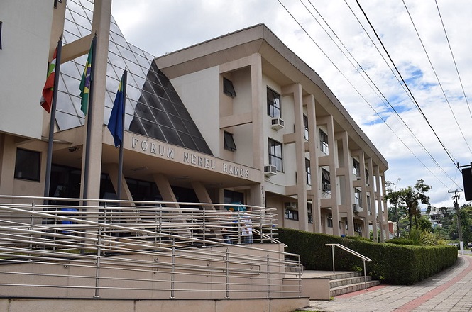 A imagem apresenta o edifício do Fórum Nereu Ramos. A fachada é composta por pilares largos e painéis de vidro inclinados que dão um ar contemporâneo ao prédio. Há uma rampa de acesso com corrimão metálico, que está em destaque na parte frontal, evidenciando acessibilidade. Na entrada, estão hasteadas bandeiras, incluindo a do Brasil. O entorno do edifício conta com áreas verdes bem cuidadas, como arbustos e árvores, o céu está parcialmente nublado.