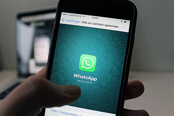 A imagem mostra um celular exibindo o aplicativo WhatsApp aberto na tela, com o logotipo centralizado em destaque. A interface está configurada em holandês, e ao fundo há um computador parcialmente visível, desfocado.