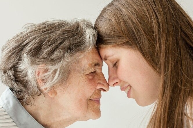 A imagem mostra uma mulher idosa e uma jovem. As duas estão de frente uma para a outra, com os rostos encostados, expressando afeição e conexão emocional. A idosa tem cabelos grisalhos curtos e um leve sorriso no rosto, enquanto a jovem, com cabelos longos castanhos, também sorri suavemente.