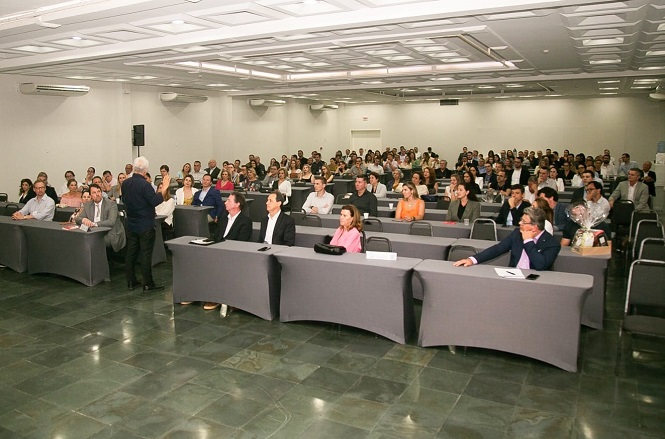 A imagem mostra uma sala de conferências cheia de pessoas. Há várias fileiras de mesas cobertas com toalhas cinza, e as pessoas estão sentadas em cadeiras atrás dessas mesas. Na frente da sala, um homem de cabelos grisalhos está em pé, aparentemente falando ou apresentando algo para o público. A sala é bem iluminada, com luzes no teto, e há um piso de azulejos escuros. O público parece estar prestando atenção ao que está sendo apresentado.