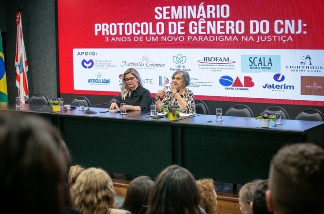 A imagem mostra uma mesa com duas palestrantes durante o Seminário “Protocolo de Gênero do CNJ: 3 anos de um novo paradigma na Justiça”. No fundo, há um painel vermelho com o título do evento e os logotipos dos apoiadores. O público está sentado de frente para as palestrantes.