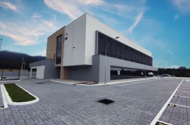 A imagem mostra o novo Fórum de Garuva. Um edifício moderno de dois andares com fachada branca e detalhes em vidro e pedra, rodeado por um estacionamento pavimentado.