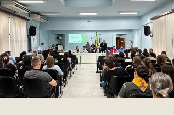 A imagem mostra uma reunião de repasse financeiro na  Comarca de São Miguel do Oeste. Em um auditório, várias pessoas estão sentadas em cadeiras voltadas para uma mesa principal, onde estão posicionadas autoridades. Ao fundo, há um projetor exibindo informações.
