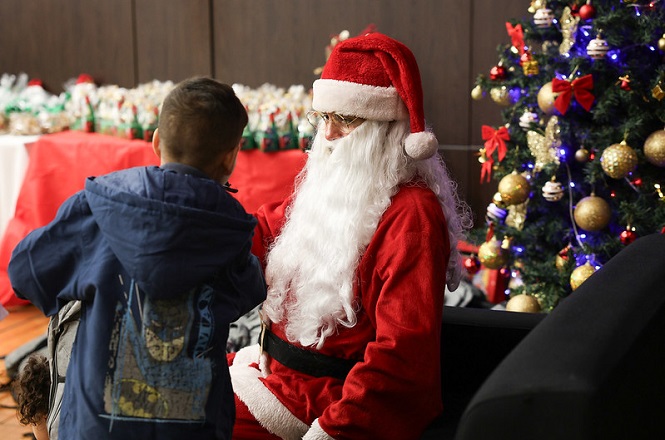 A imagem mostra uma cena natalina com o Papai Noel sentado em uma cadeira. Ele está vestido com o tradicional traje vermelho e branco, incluindo o chapéu e a barba longa e branca. À sua frente, há uma criança de costas, usando uma jaqueta azul escura. Ao lado direito da imagem, há uma árvore de Natal decorada com bolas douradas e vermelhas, além de luzes coloridas. No fundo, há uma mesa coberta com uma toalha vermelha, cheia de sacolas ou presentes embrulhados. A atmosfera é festiva e alegre, típica de uma celebração de Natal.