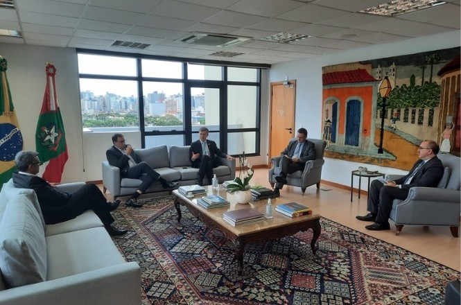A imagem mostra uma sala de reuniões com cinco homens sentados em sofás e poltronas dispostas em um formato de círculo. Dentre eles estão o presidente do TJSC, desembargador Francisco Oliveira Neto, e o presidente da Fecam, Kleber Wan-Dall, prefeito de Gaspar. A sala é bem iluminada, com uma grande janela ao fundo que oferece uma vista da cidade. No centro da sala, há uma mesa de centro de madeira com alguns livros e um vaso de plantas. No canto esquerdo, há duas bandeiras, uma do Brasil e outra de Santa Catarina.