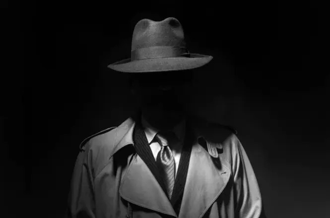 A imagem mostra uma figura masculina em um ambiente escuro, vestindo um sobretudo e um chapéu de feltro, típico de detetives dos anos 1940 ou 1950. A iluminação é baixa e cria um efeito de sombra que cobre parcialmente o rosto do homem, dando um ar de mistério. Ele está usando uma gravata e o colarinho da camisa está visível. A cena tem um estilo noir, evocando filmes de suspense ou investigação daquela época.