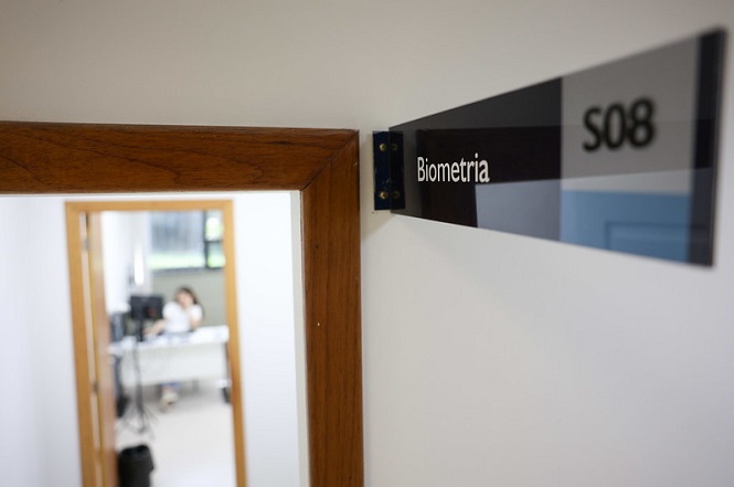 A imagem mostra a entrada de uma sala com uma placa na parede ao lado da porta. A placa tem a palavra "Biometria" escrita e o número "S08". A porta tem uma moldura de madeira e, através dela, é possível ver uma pessoa ao fundo, sentada em uma mesa, embora a imagem esteja um pouco desfocada nessa parte. A sala parece ser bem iluminada.