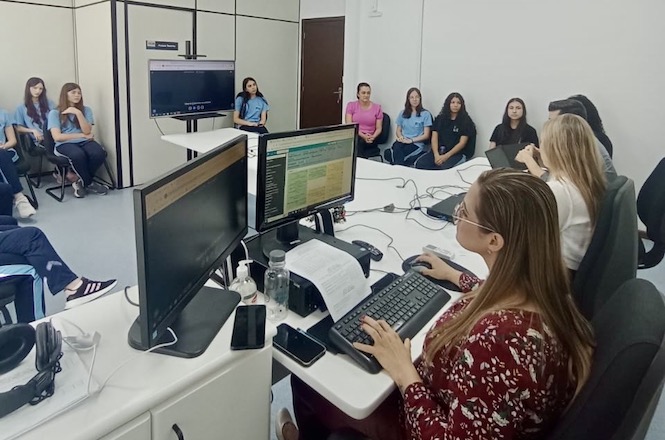 A imagem mostra uma sala de reunião onde várias pessoas estão reunidas. No primeiro plano, há uma mulher de cabelos longos e castanhos, usando óculos e uma blusa vermelha com estampas florais, sentada em frente a dois monitores de computador. Ela está digitando em um teclado. Ao redor da mesa, há outras pessoas sentadas, algumas delas olhando para os monitores. No fundo da sala, há um grupo de mulheres sentadas em cadeiras contra a parede, algumas vestindo camisetas azuis e outras de preto. Na parede à esquerda, há uma tela grande, possivelmente uma TV ou monitor, que parece estar ligada. A sala tem paredes brancas e um ambiente de escritório.