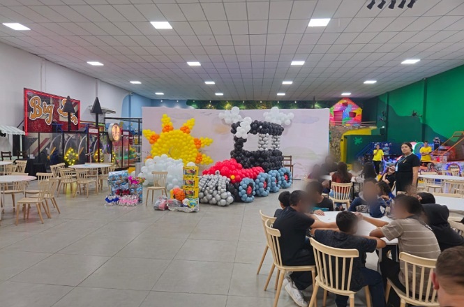 A imagem mostra um salão de festas decorado para uma celebração infantil. No centro, há uma grande escultura de balões em forma de trem, com balões pretos, vermelhos, cinzas e brancos. Ao lado do trem, há uma representação do sol feita com balões amarelos e laranjas. Em volta, há várias mesas e cadeiras de madeira clara, algumas ocupadas por pessoas. No fundo, há brinquedos e estruturas coloridas, típicas de um espaço de recreação infantil. As paredes são brancas e o teto é coberto por placas de iluminação.