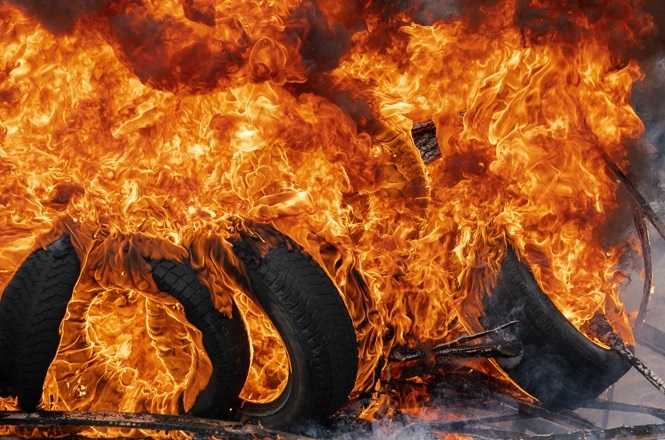 A imagem mostra pneus em chamas. As chamas são intensas e cobrem quase toda a cena, com cores vibrantes de laranja e amarelo. Os pneus estão parcialmente visíveis no meio do fogo, e a intensidade das chamas sugere um incêndio de grandes proporções. A cena transmite uma sensação de calor e destruição.