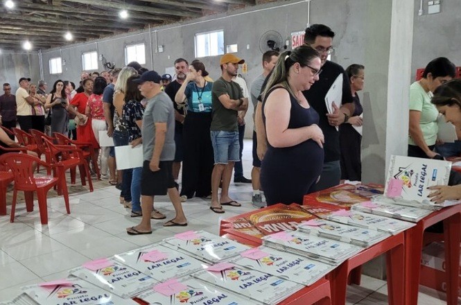 A imagem mostra uma fila de pessoas em um ambiente interno, que parece ser um salão ou auditório. As pessoas estão em pé, aguardando em uma fila que se estende ao longo do salão. À direita da imagem, há uma mesa vermelha com vários pacotes ou pastas brancas sobre ela, cada uma com um adesivo rosa. Em algumas dessas pastas está escrito "LAR LEGAL". As pessoas na fila seguram papéis ou documentos. O ambiente tem cadeiras plásticas vermelhas empilhadas à esquerda e um teto com iluminação. As paredes são de concreto e há algumas janelas pequenas.