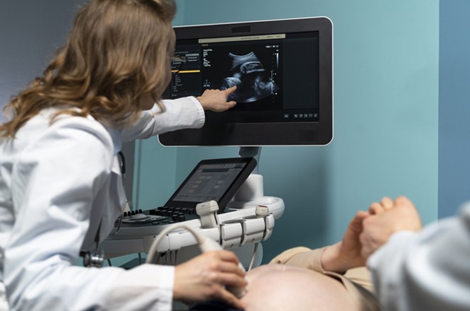 A imagem mostra uma médica realizando um exame de ultrassom em uma paciente grávida. A médica está usando um jaleco branco e está apontando para a tela do monitor, que exibe a imagem do ultrassom. A paciente está deitada, com a barriga exposta, enquanto a médica utiliza o transdutor do ultrassom sobre a barriga da paciente. O ambiente parece ser uma sala de exames com paredes de cor azul clara.
