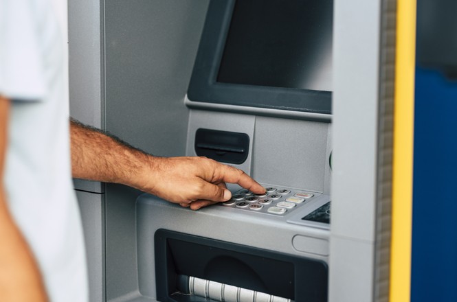 A imagem mostra uma pessoa usando um caixa eletrônico. A mão da pessoa está pressionando os botões do teclado numérico do caixa. O caixa eletrônico tem uma tela acima do teclado e uma abertura abaixo, que é onde o dinheiro ou recibos são dispensados. A pessoa está posicionada à esquerda da imagem, e apenas parte do braço e da mão são visíveis.