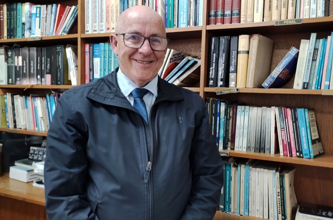A imagem mostra um homem idoso sorrindo, usando óculos, uma jaqueta escura e uma gravata azul clara com camisa branca. Ele está em pé em frente a uma estante cheia de livros. A estante é de madeira e contém muitos livros de diferentes tamanhos e cores, organizados em várias prateleiras. O ambiente parece ser um escritório ou uma biblioteca.