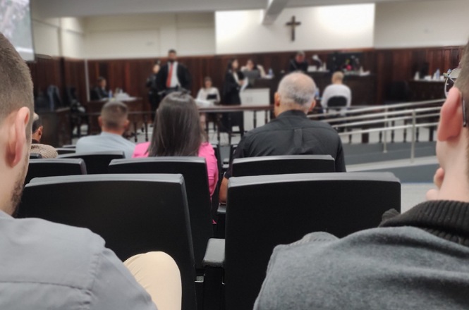 A imagem mostra o interior de uma sala de tribunal. No primeiro plano, há várias pessoas sentadas em cadeiras, vistas de costas. No fundo, há um grupo de pessoas em pé e sentadas atrás de uma bancada elevada, possivelmente juízes e advogados. A sala tem um crucifixo na parede acima da bancada, indicando que pode ser um tribunal em um país de tradição cristã. As pessoas na bancada estão vestidas formalmente, e uma delas parece estar falando ou apresentando algo. A iluminação é clara e o ambiente parece ser sério e formal.