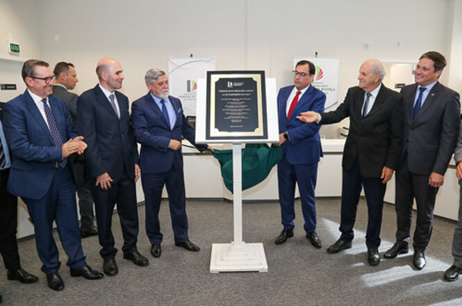 A imagem mostra um grupo de sete homens em pé, todos vestindo ternos, em um evento de inauguração ou cerimônia oficial. No centro da imagem, há uma placa em um suporte, que parece ser o foco da cerimônia. Dois dos homens estão segurando uma bandeira verde que foi removida para revelar a placa. Atrás do grupo, há dois banners com logotipos que parecem ser de uma instituição ou organização. O ambiente é bem iluminado e parece ser uma sala de conferências ou um espaço similar.