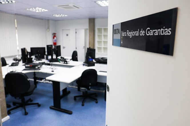 A imagem mostra uma sala de escritório com várias mesas e cadeiras. As mesas estão equipadas com computadores e monitores. A sala tem piso azul e iluminação de teto com lâmpadas fluorescentes. Na porta, há uma placa com o texto "007A Vara Regional de Garantias". Ao fundo, é possível ver uma parede com janelas cobertas por persianas e uma porta de saída de emergência com um extintor ao lado.