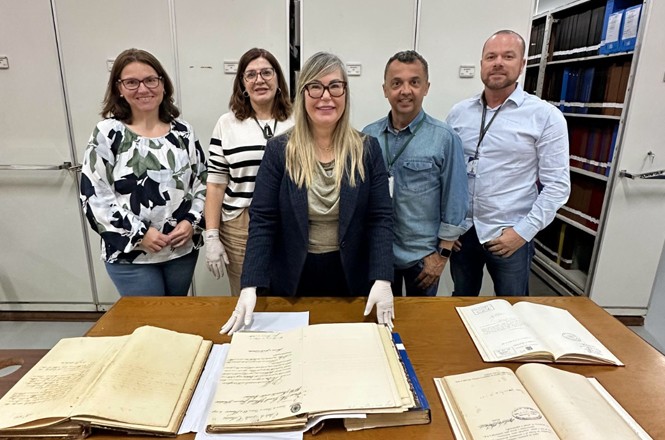 A imagem mostra cinco pessoas em pé atrás de uma mesa de madeira, que está coberta com livros e documentos antigos abertos. As pessoas estão em um ambiente que parece ser um arquivo ou biblioteca, com estantes cheias de livros ao fundo. Três mulheres e dois homens estão na foto. A mulher no centro é a desembargadora Haidée Denise Grin que está usando óculos e luvas brancas, assim como a mulher à sua esquerda. Todos estão sorrindo e olhando para a câmera. As estantes ao fundo são de metal e estão cheias de livros encadernados.