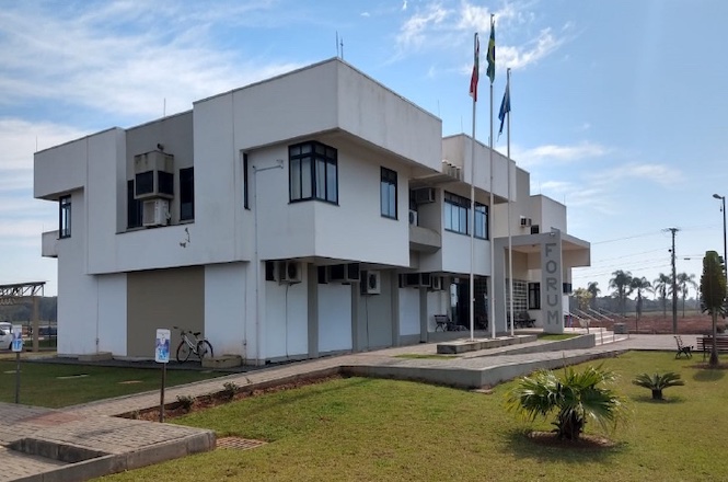 A imagem mostra um edifício de dois andares com arquitetura moderna e linhas retas, identificado como um fórum. Na fachada, há a palavra "FÓRUM" escrita verticalmente em uma estrutura próxima à entrada principal. O prédio é branco e possui várias janelas de vidro. Na frente do edifício, há um gramado bem cuidado com algumas pequenas plantas. Três mastros com bandeiras estão posicionados ao lado da entrada, e as bandeiras estão hasteadas. O céu está parcialmente nublado, e o ambiente parece ser de um dia claro. Há também uma bicicleta estacionada próxima à entrada do prédio.