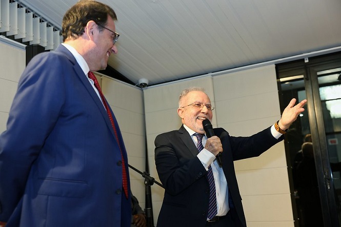 A imagem mostra o Presidente do TJSC, des. Francisco Oliveira Neto, à esquerda, vestindo um terno azul, camisa branca e gravata vermelha e o desembargador Pedro Manoel Abreu à direita, usando óculos e um terno preto com camisa branca e gravata listrada. Ele está segurando um microfone e parece estar falando ou fazendo um discurso, com uma expressão sorridente e um braço levantado. O ambiente parece ser uma sala de eventos ou conferências, com paredes claras e uma porta de vidro ao fundo.