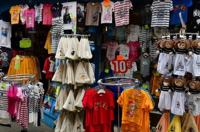 A imagem mostra uma loja de roupas infantis, com várias peças penduradas e expostas. Há camisetas de várias cores e estampas, incluindo listradas, com personagens de desenhos animados e outras ilustrações. No centro, há um suporte com várias capas ou ponchos infantis pendurados, de cor clara com pequenos desenhos. À direita, há uma fileira de macacões infantis com capuz, decorados com rostos de macacos.