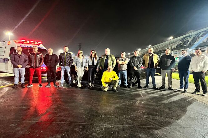 A imagem mostra um grupo de pessoas posando para uma foto em um ambiente externo à noite. Elas estão alinhadas em pé, com exceção de uma pessoa que está agachada na frente do grupo. Ao fundo, à esquerda, há uma ambulância com as luzes vermelhas acesas. O local parece ser um campo de futebol. As pessoas estão vestidas de maneira casual, com jaquetas e calças, e o clima parece estar frio.
