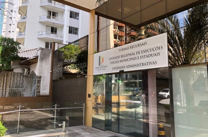 A imagem mostra a fachada de um prédio com uma placa na entrada. A placa tem o logotipo do Poder Judiciário do Estado do Espírito Santo e os seguintes dizeres: "TURMAS RECURSAIS UNIDADE REGIONAL DE EXECUÇÕES FISCAIS MUNICIPAIS E ESTADUAIS UNIDADES ADMINISTRATIVAS". O prédio é moderno, com paredes de vidro e um corrimão metálico na entrada. Ao fundo, é possível ver outros edifícios residenciais e algumas árvores.