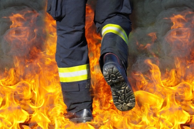 A imagem mostra as pernas de uma pessoa vestindo uma calça de bombeiro, que é de cor escura com faixas refletivas amarelas. A pessoa está caminhando em meio a chamas intensas, que cercam seus pés e pernas. As botas são robustas e parecem ser feitas de um material resistente ao fogo.