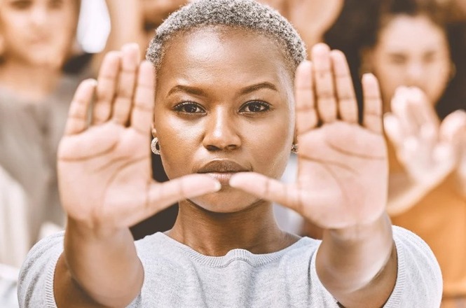A imagem mostra uma mulher negra de cabelo curto e grisalho, olhando diretamente para a câmera. Ela está com as mãos levantadas à frente do rosto, com as palmas abertas e voltadas para fora, como se estivesse fazendo um gesto de "pare". Ao fundo, há outras pessoas, mas estão desfocadas, destacando a mulher em primeiro plano. A expressão dela é séria e determinada.