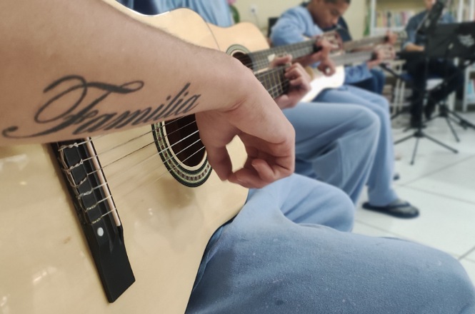 A imagem mostra uma pessoa tocando violão. O foco está na mão direita da pessoa, que está dedilhando as cordas do violão. Ao fundo, é possível ver outras pessoas também tocando violão, todas vestidas com roupas azuis. A cena parece ser em um ambiente interno, um espaço de prática musical.