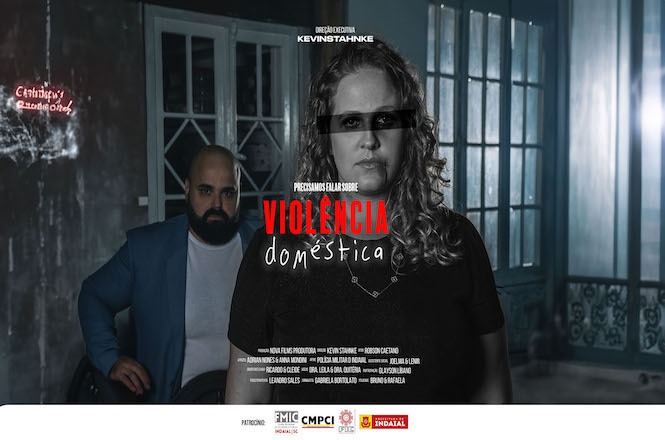 A imagem é um cartaz de um filme com o título "Precisamos falar sobre Violência Doméstica". No centro do cartaz, há uma mulher de cabelos loiros encaracolados, com uma expressão séria. Seus olhos estão cobertos por uma faixa preta, simbolizando anonimato ou censura. Atrás dela, há um homem careca com barba, vestindo um paletó azul e uma camiseta branca, sentado em uma cadeira de rodas. O ambiente parece ser uma sala com janelas grandes ao fundo. No canto superior esquerdo, há um texto em vermelho que diz "Catubiry Studio One". A direção é de Kevin Stahnke. Na parte inferior do cartaz, há logotipos de patrocinadores, incluindo FMIC, CMPCI, Prefeitura de Caxias do Sul e Indavideo.
