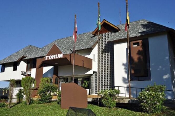 A imagem mostra um edifício identificado como "FÓRUM", com a palavra escrita em letras brancas em uma placa marrom acima da entrada principal. O prédio tem um design moderno com telhado inclinado coberto por telhas cinzas. Há três mastros com bandeiras na frente do edifício, e o ambiente ao redor é bem cuidado, com gramado e arbustos. O céu está claro e azul, indicando que é um dia ensolarado.