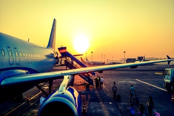 A imagem mostra um avião estacionado em um aeroporto durante o pôr do sol. O sol está baixo no horizonte, criando um céu alaranjado e iluminando a cena com uma luz dourada. À esquerda, vê-se a fuselagem do avião, com suas janelas visíveis. Há uma escada móvel encostada na porta do avião, permitindo o embarque e desembarque de passageiros. No lado direito da imagem, algumas pessoas estão caminhando em direção ao avião, carregando malas. Ao fundo, é possível ver outros elementos do aeroporto, como veículos de serviço e estruturas de apoio.