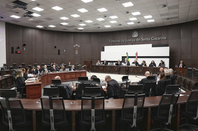 A imagem mostra uma sala de tribunal do Tribunal de Justiça de Santa Catarina. A sala é ampla, com um teto alto e várias luzes embutidas. As paredes são revestidas de madeira escura. No centro da imagem, há uma grande mesa em formato de U, onde várias pessoas estão sentadas, todas vestidas com togas pretas, típicas de juízes e advogados. Na parede ao fundo, está escrito "Tribunal de Justiça de Santa Catarina" e há um brasão acima do texto. Também há um crucifixo pendurado na parede à esquerda. No centro da mesa principal, há bandeiras do Brasil e de Santa Catarina. A sala está organizada de forma a acomodar muitas pessoas, com várias cadeiras dispostas ao redor.
