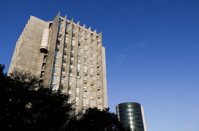 A imagem mostra dois edifícios sob um céu azul claro. O edifício à esquerda é alto com concreto aparente e várias janelas verticais. O edifício à direita está em segundo plano, tem uma forma cilíndrica, com janelas espelhadas que refletem a luz. Na parte inferior da imagem, há algumas árvores que parcialmente obscurecem a base dos edifícios.