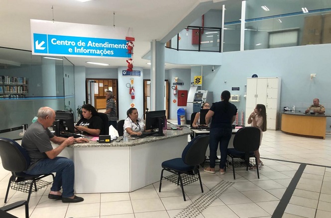 A imagem mostra um ambiente interno de um centro de atendimento e informações. No centro da imagem, há um balcão em formato de meia-lua com pessoas sentadas em frente a computadores, atendendo o público. No fundo, há um letreiro azul com a inscrição "Centro de Atendimento e Informações". O ambiente é bem iluminado e possui piso claro.