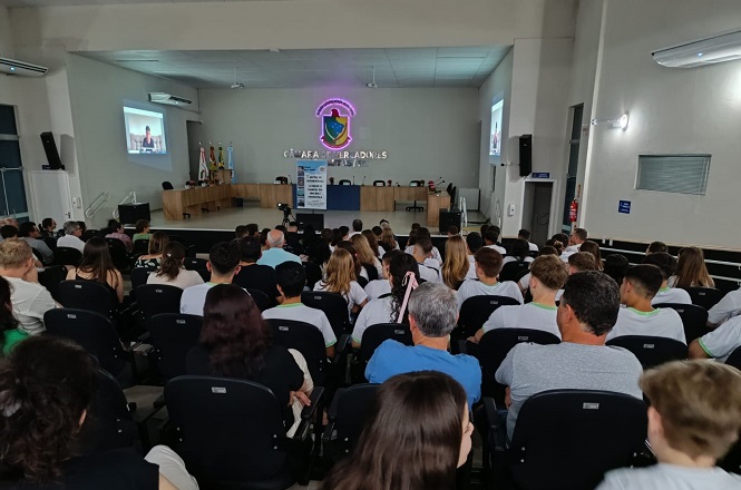 A imagem mostra uma sala de conferências ou auditório com várias pessoas sentadas, olhando para a frente. Na parte frontal da sala, há um palco com uma mesa comprida e várias cadeiras. Atrás da mesa, na parede, está o brasão da cidade e as palavras "Câmara de Vereadores". Há também um púlpito com um banner ao lado. Nas laterais da sala, há projetores exibindo imagens nas paredes. As pessoas na audiência parecem estar assistindo a uma apresentação ou reunião. O ambiente é bem iluminado e organizado.