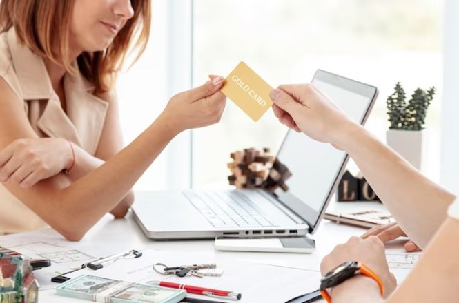 A imagem mostra duas pessoas em um ambiente de escritório. Uma mulher, à esquerda, está sentada à mesa, vestindo uma blusa sem mangas bege. Ela está recebendo um cartão dourado com a inscrição "GOLD CARD" de uma pessoa à direita, cuja mão está estendida. Na mesa, há um laptop aberto, algumas chaves, notas de dinheiro, uma caneta vermelha e alguns papéis. Ao fundo, há uma pequena planta em um vaso branco e um objeto decorativo. A cena parece retratar uma transação ou uma troca de informações financeiras.