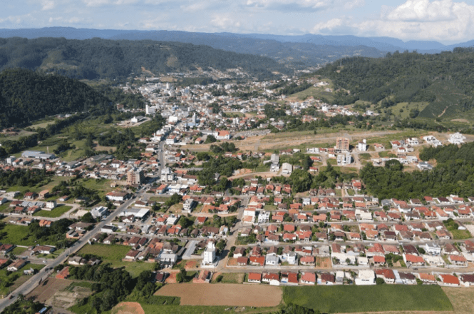 A imagem mostra uma vista aérea de uma cidade cercada por colinas e áreas verdes. A cidade possui várias casas e edifícios, com ruas organizadas em um padrão de grade. As construções são predominantemente de pequeno a médio porte, com telhados vermelhos e algumas áreas de vegetação entre as casas. Ao fundo, é possível ver colinas cobertas de vegetação, e o céu está parcialmente nublado. A cidade parece estar em uma área montanhosa, com um relevo variado ao redor.
