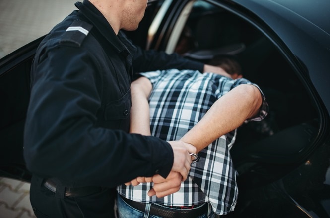 A imagem mostra um policial prendendo um homem. O policial está vestindo um uniforme preto e está algemando o homem, que está inclinado para frente, com as mãos para trás. O homem está vestindo uma camisa xadrez em tons de preto e branco e jeans. Eles estão ao lado de um carro preto, e parece que o homem está sendo colocado dentro do veículo. A cena ocorre em um ambiente externo, possivelmente em um estacionamento.