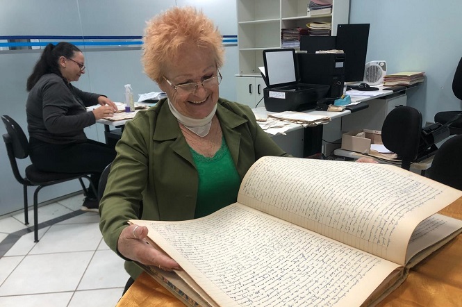 A imagem mostra uma mulher idosa sorridente, com cabelo curto e ruivo, usando óculos e vestindo uma blusa verde com um casaco verde-oliva por cima. Ela está sentada em uma mesa e segurando um grande livro antigo com páginas cheias de escrita manuscrita. Ao fundo, há outra mulher sentada em uma mesa, ocupada com papéis e um computador.