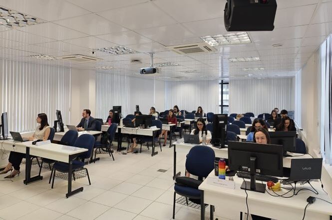 A imagem mostra uma sala com várias pessoas sentadas em frente a computadores. As mesas estão dispostas em fileiras, e cada uma tem um ou dois computadores. As pessoas estão concentradas em suas telas. A sala é bem iluminada, com luz natural entrando pelas janelas, que têm persianas verticais brancas. As paredes são brancas, e o piso é de cerâmica clara. Há também um ar-condicionado instalado no teto.