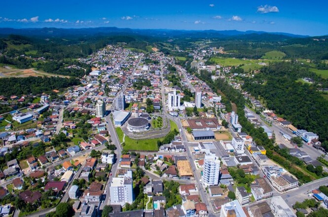 A imagem mostra uma vista aérea de uma cidade. No centro da imagem, há um edifício circular grande, cercado por ruas e outros prédios. A cidade parece ser de médio porte, com várias casas e edifícios espalhados. Há algumas construções mais altas, principalmente no centro da imagem. A área ao redor da cidade é verde, com colinas e vegetação densa. O céu está azul com algumas nuvens dispersas, indicando um dia claro e ensolarado. A cidade está situada em um vale, cercada por colinas ao fundo.